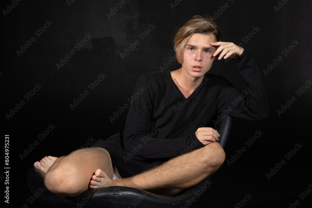 Fototapeta premium A young attractive guy is sad while sitting on a chair. Black background.