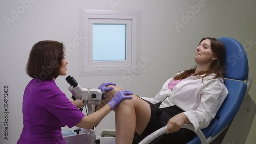A female patient smiles and closes her eyes in fear, while sitting in a gynecological chair before a colposcopy procedure. A female gynecologist in a purple medical suit is trying to calm her client photo