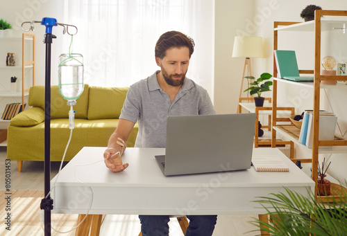 Young man sitting and working on a laptop at the desk of his workplace at home or in office while receiving IV drip infusion and vitamin therapy in his blood. Male person receiving injection therapy.