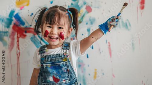 A cheerful mixed-race girl wearing overalls is depicted holding an artist's paintbrush, with her hands raised high and splashes of dark red color covering her from painting the wall. 
