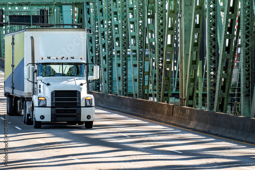 White day cab big rig semi truck with roof spoiler transporting cargo in dry van semi trailer running on the truss arched bridge