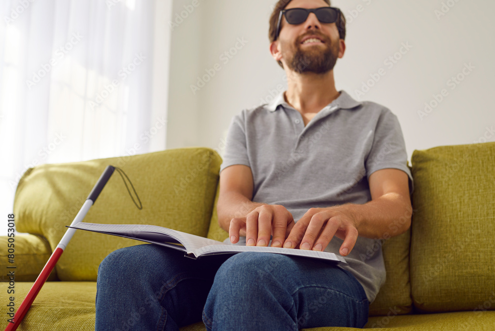 Portrait of a smiling bearded blind man in black eyeglasses sitting on sofa in the living room at home with stick cane and reading tactile braille book. Blindness and education concept. Portrait of a smiling bearded blind man in black eyeglasses sitting on sofa in the living room at home with stick cane and reading tactile braille book. Blindness and education concept.