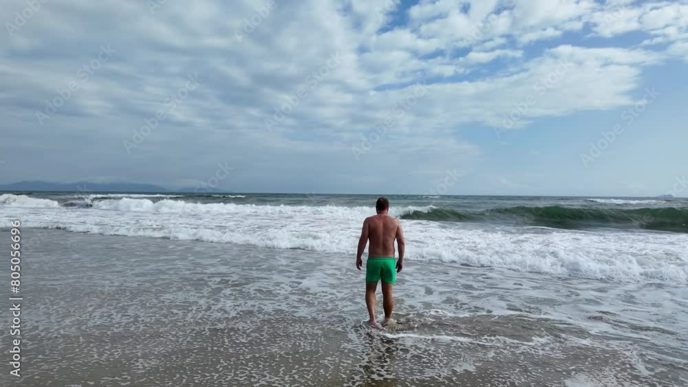 Back view of an adult male in green shorts standing by the sea on a sunny beach day, concept of summer vacation and relaxation Back view of an adult male in green shorts standing by the sea on a sunny beach day, concept of summer vacation and relaxation