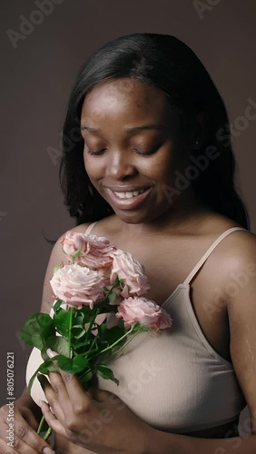 Vertical studio shot of young beautiful Black woman in minimalist bra smelling fresh roses and happily smiling