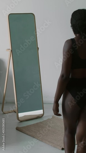 Vertical shot of pretty Black girl in underwear entering bedroom and looking at her body in mirror with love