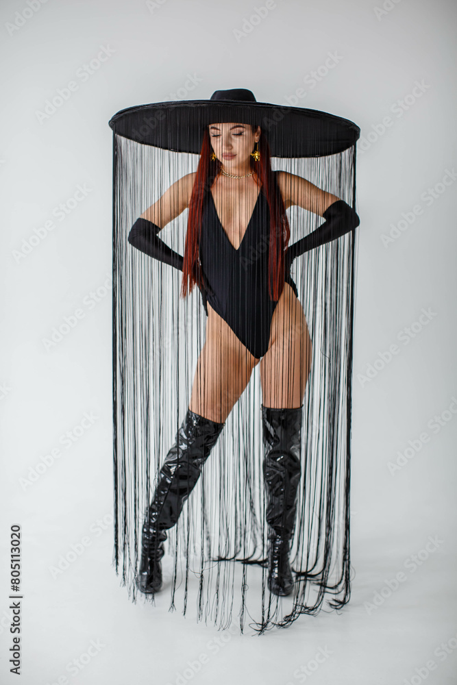 A red-haired girl in a black bodysuit, patent leather boots, gloves and a hat with threads stands in the studio on a white background