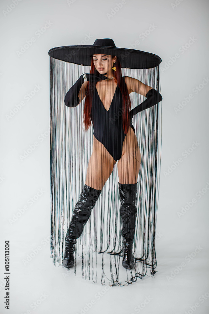 A red-haired girl in a black bodysuit, patent leather boots, gloves and a hat with threads stands in the studio on a white background