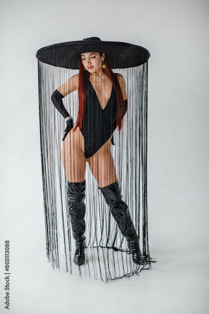 A red-haired girl in a black bodysuit, patent leather boots, gloves and a hat with threads stands in the studio on a white background