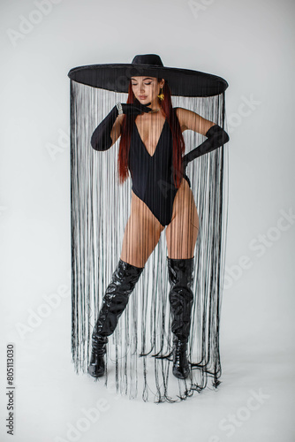 Obraz na płótnie A red-haired girl in a black bodysuit, patent leather boots, gloves and a hat with threads stands in the studio on a white background w salonie
