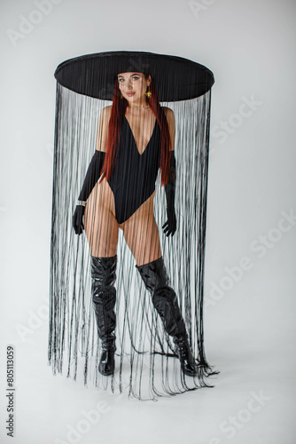 A red-haired girl in a black bodysuit, patent leather boots, gloves and a hat with threads stands in the studio on a white background
