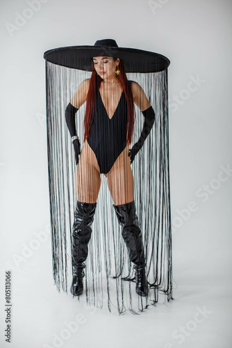 Obraz na płótnie A red-haired girl in a black bodysuit, patent leather boots, gloves and a hat with threads stands in the studio on a white background w salonie