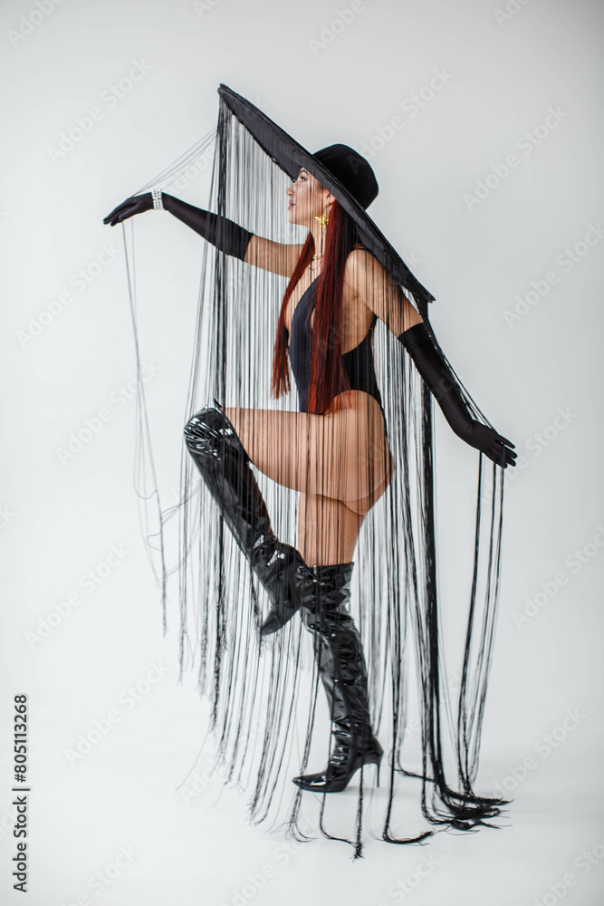 A red-haired girl in a black bodysuit, patent leather boots, gloves and a hat with threads stands in the studio on a white background