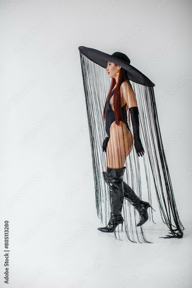 A red-haired girl in a black bodysuit, patent leather boots, gloves and a hat with threads stands in the studio on a white background