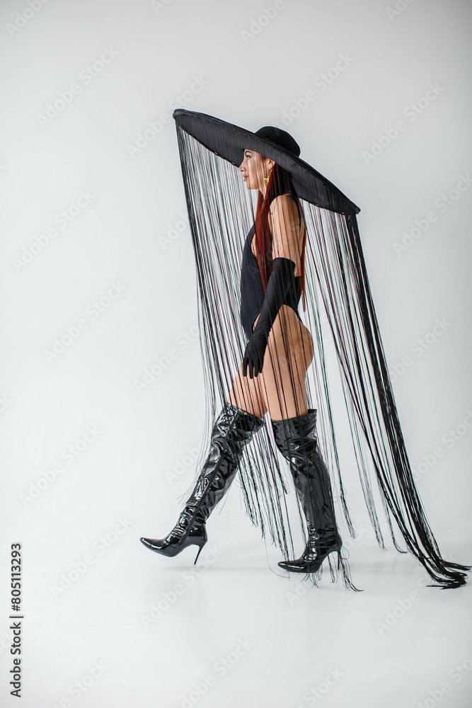 A red-haired girl in a black bodysuit, patent leather boots, gloves and a hat with threads stands in the studio on a white background