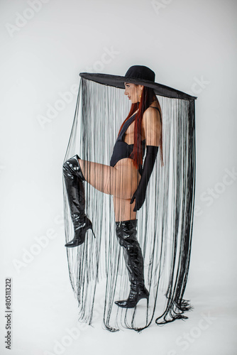 A red-haired girl in a black bodysuit, patent leather boots, gloves and a hat with threads stands in the studio on a white background