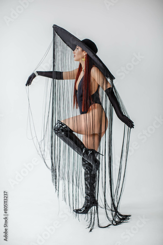 Obraz na płótnie A red-haired girl in a black bodysuit, patent leather boots, gloves and a hat with threads stands in the studio on a white background w salonie