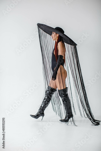 Obraz na płótnie A red-haired girl in a black bodysuit, patent leather boots, gloves and a hat with threads stands in the studio on a white background w salonie