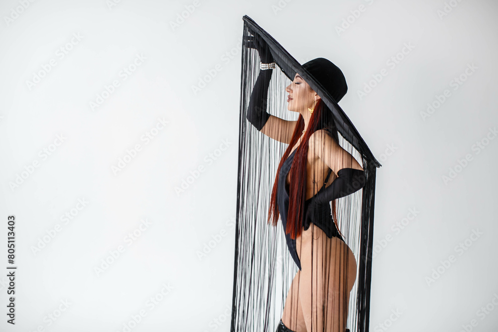 A portrait of a red-haired girl in a black bodysuit, gloves and a hat with threads stands in the studio on a white background.