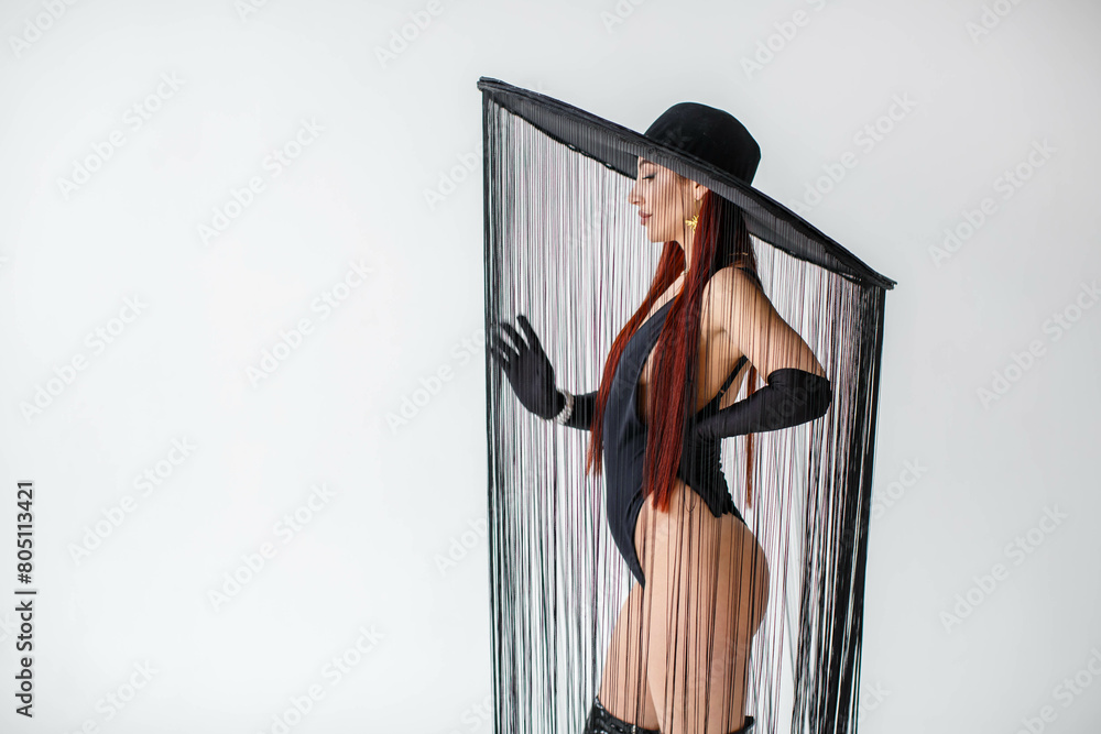 A portrait of a red-haired girl in a black bodysuit, gloves and a hat with threads stands in the studio on a white background.