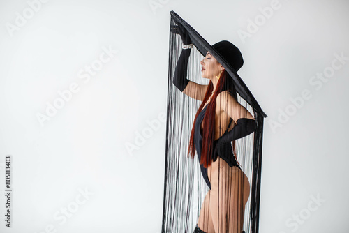 A portrait of a red-haired girl in a black bodysuit, gloves and a hat with threads stands in the studio on a white background.