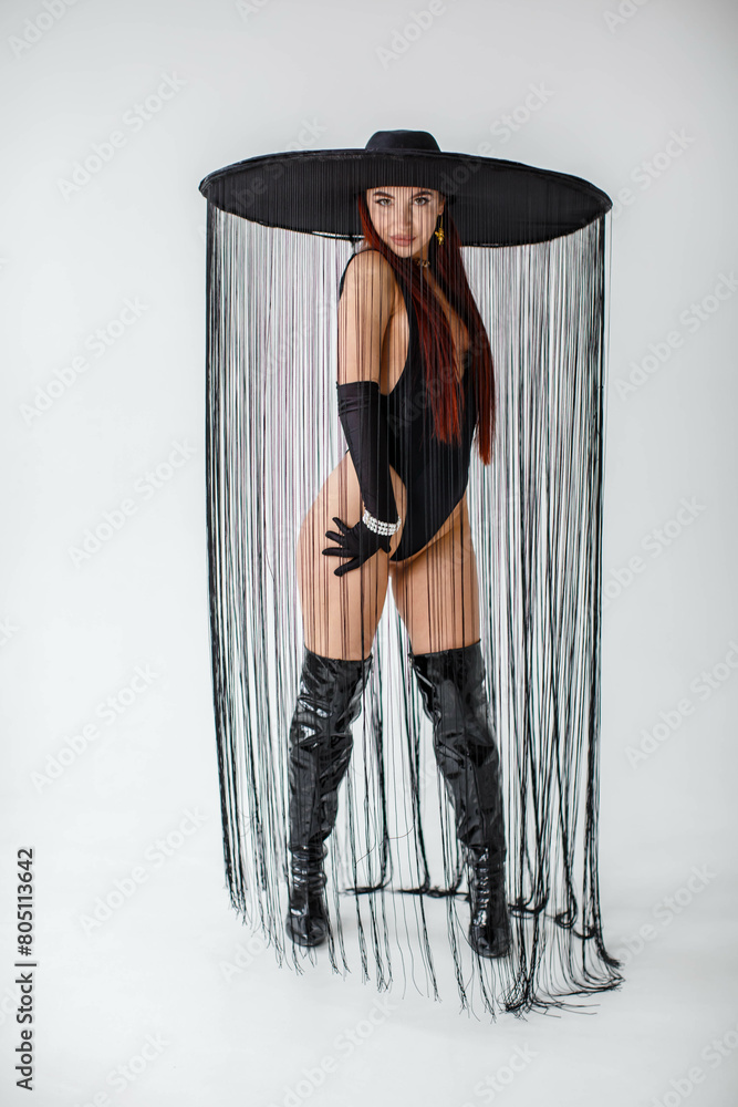 girl in a black bodysuit, patent leather boots, gloves and a hat with threads in the studio on a white background