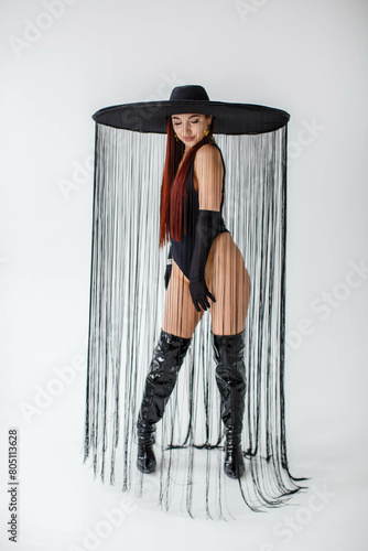 A red-haired girl in a black bodysuit, patent leather boots, gloves and a hat with threads stands in the studio on a white background