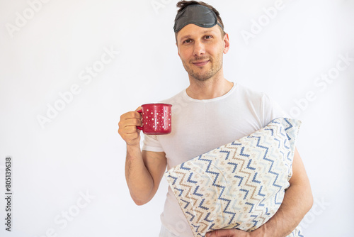 portrait of a young man in pajamas holding a pillow and a cup isolated on white background