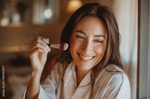 A beautiful woman in her bathroom  smiling and applying powder with the brush to one side of face  wearing white robe