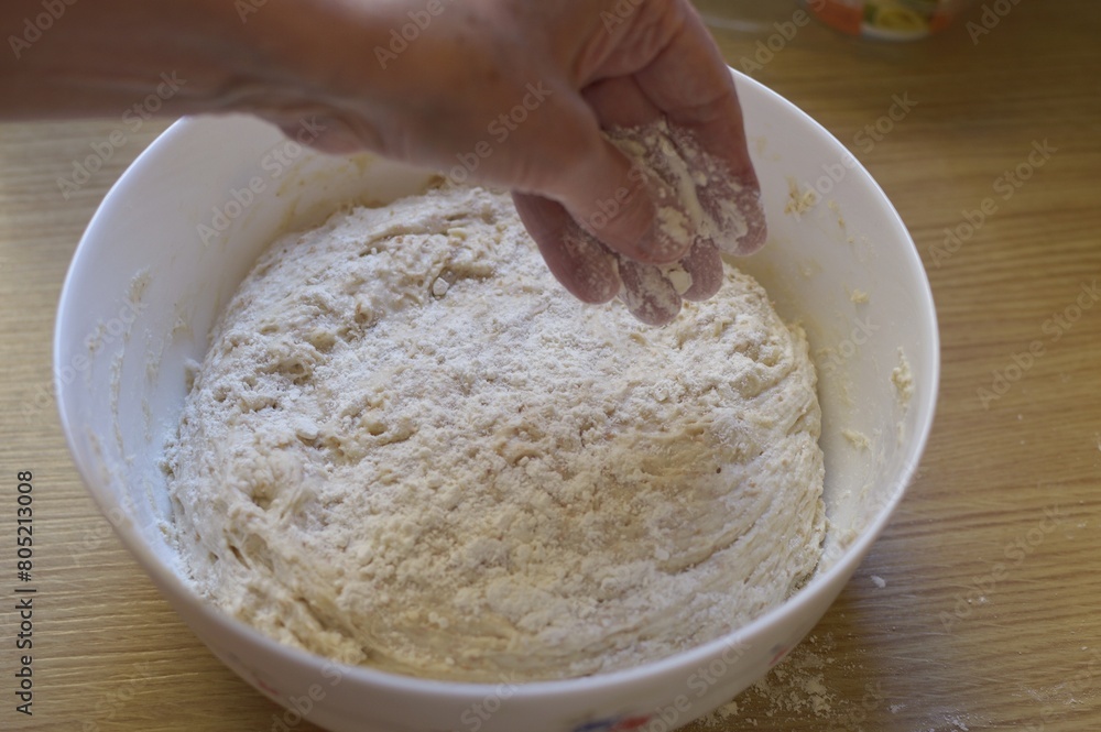 hand adds flour onto raw yeast dough in a ceramic bowl on a wooden kitchen table. hand adds flour onto raw yeast dough in a ceramic bowl on a wooden kitchen table.