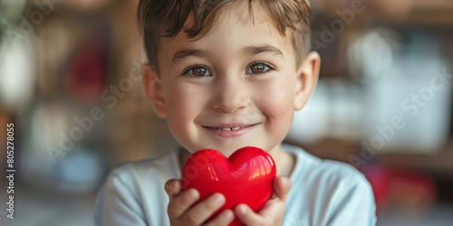 A boy holding a red heart shape and smiling at the camera in a close-up portrait of a happy child with a big love symbol.
