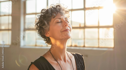 An attractive woman with short curly hair and closed eyes stands in a yoga studio, engaging in deep breathing exercises while looking upward. photo