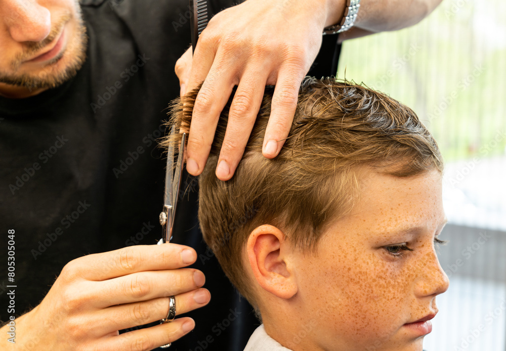 Fototapeta premium Children hairdresser with scissors is cutting little boy against a dark background. Contented cute preschooler boy getting the haircut. 