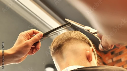 Man barber cutting little boy's hair using comb and scissors. Child getting haircut from adult male, likely barber. Professional hairdresser and cute client at modern barbershop. photo