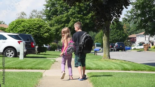 Two young children are wearing book bags and walking to school on a suburban sidewalk outside on a sunny day with backpacks for an back-to-school or safety concept.