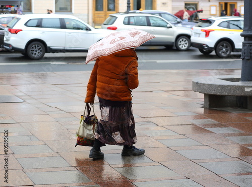 An old woman with an umbrella walks in the rain on wet granite slabs of the sidewalk, Nevsky Prospekt, St. Petersburg, Russia, May 04, 2024