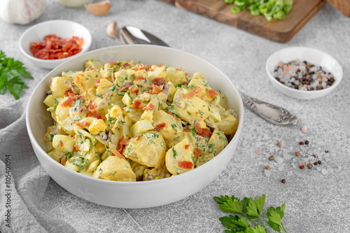 Salad of potatoes, eggs, bacon and fresh herbs with mayonnaise and yogurt dressing in a bowl on a gray concrete background.