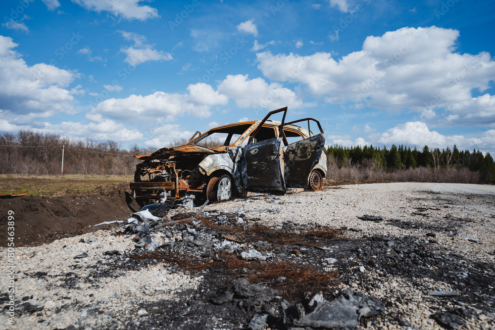 A burnt car is abandoned on the road in a field against the background of the sky, rusty iron, charred earth.