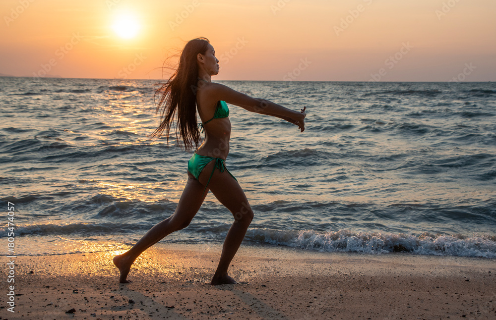 Fototapeta premium Asian tourist wearing a sexy bikini, exercising, stretching, doing yoga, beautiful beach by the sea, sunset, relaxing, summer time, vacation.