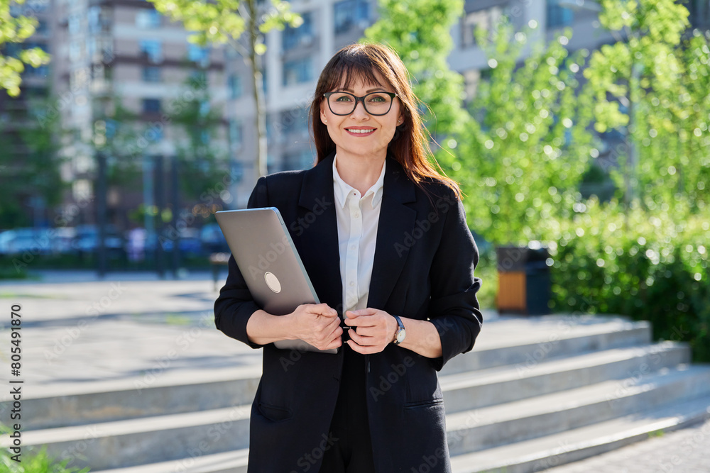 Middle-aged business woman in suit with laptop, outdoor near building Middle-aged business woman in suit with laptop, outdoor near building