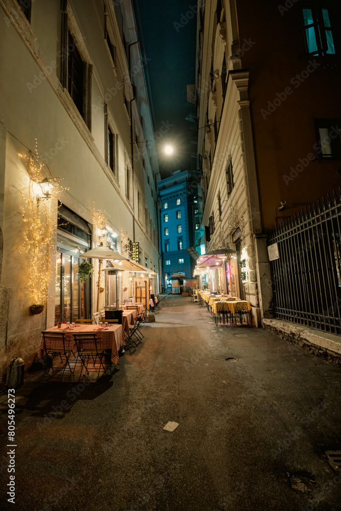 Fototapeta premium In a quaint narrow street at night, tables and chairs are set up under the dim glow of streetlights. Customers enjoy dining, chatting, and sipping drinks al fresco