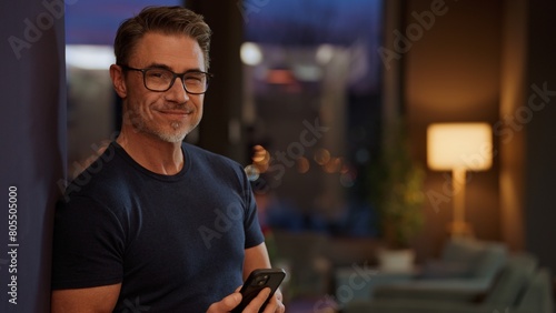 Casual mid adult man with cell phone at home. Home office, banking online, checking social media. Portrait of older bearded guy smiling. Using mobile application, telecommunication.
