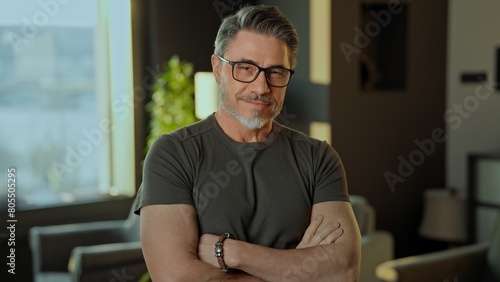 Smiling happy man in his 50s with crossed arms standing in a modern living room. Portrait of confident fit muscular mid adult older male in 50s wearing glasses. © nyul
