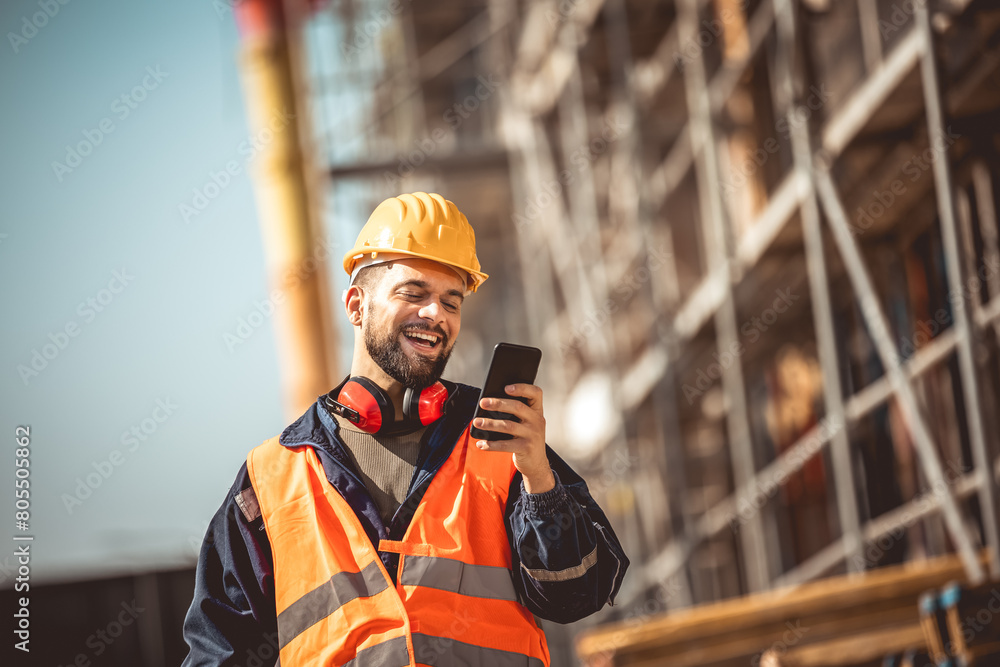 Construction site manager standing wearing safety vest and helmet,using his phone at construction site. Construction site manager standing wearing safety vest and helmet,using his phone at construction site.