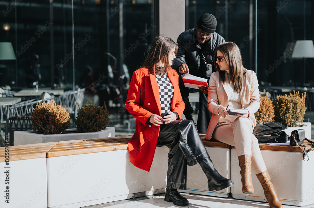 Two women in stylish outerwear are engaged in a casual business discussion while seated outdoors, with a male colleague standing over them. Two women in stylish outerwear are engaged in a casual business discussion while seated outdoors, with a male colleague standing over them.