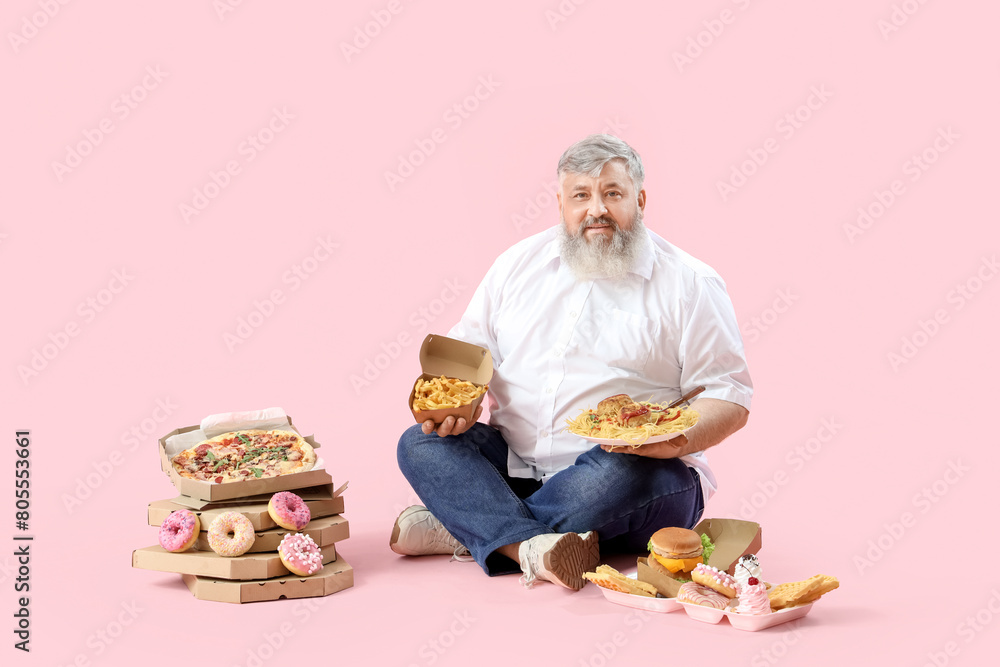 Overweight mature man with unhealthy food sitting on pink background. Overeating concept Overweight mature man with unhealthy food sitting on pink background. Overeating concept