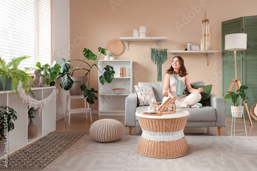 Foto Young woman meditating on sofa and hanging wind chime in living room