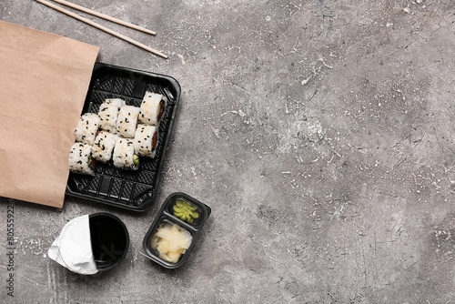 Plastic plate with tasty sushi rolls, chopsticks and paper bag on grey grunge background. Delivery concept