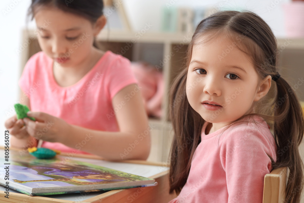 Cute little Asian happy sisters with toys reading book at table in child room Cute little Asian happy sisters with toys reading book at table in child room