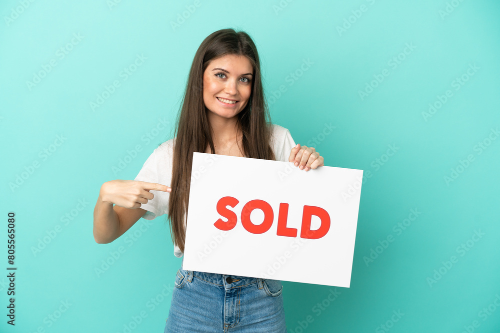 Young caucasian woman isolated on blue background holding a placard with text SOLD and pointing it Young caucasian woman isolated on blue background holding a placard with text SOLD and pointing it