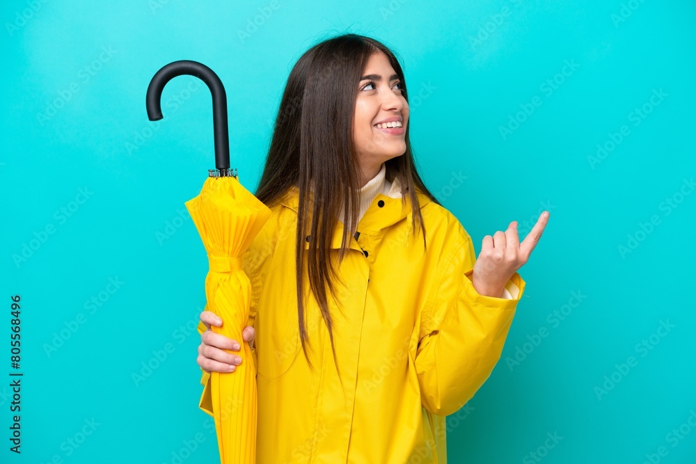 Young caucasian woman with rainproof coat and umbrella isolated on blue background pointing up a great idea Young caucasian woman with rainproof coat and umbrella isolated on blue background pointing up a great idea